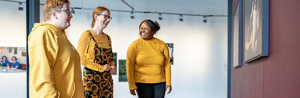 Bidh buidheann dhaoine a’ gàireachdainn agus iad a’ faighinn tlachd à taisbeanadh dhealbhan ann an gailearaidh ealain.