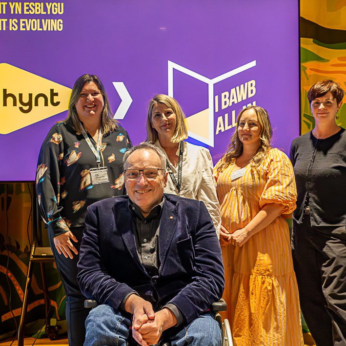 Four white people standing in front of a screen showing the Hynt and All in logos. One person in the front is wearing glasses and is in a wheelchair.