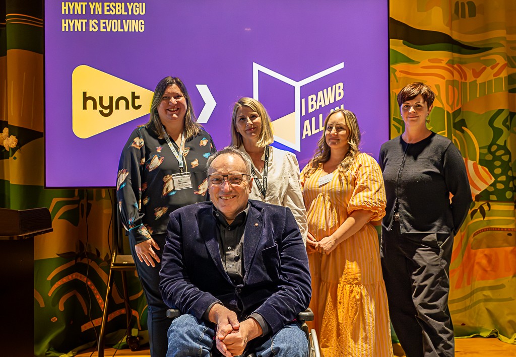 Four white people standing in front of a screen showing the Hynt and All in logos. One person in the front is wearing glasses and is in a wheelchair.